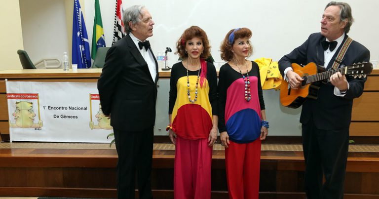 Dois homens de terno, gêmeos, em um palco, um deles com violão, e duas meninas gêmeas com roupas iguais, de cores diferentes