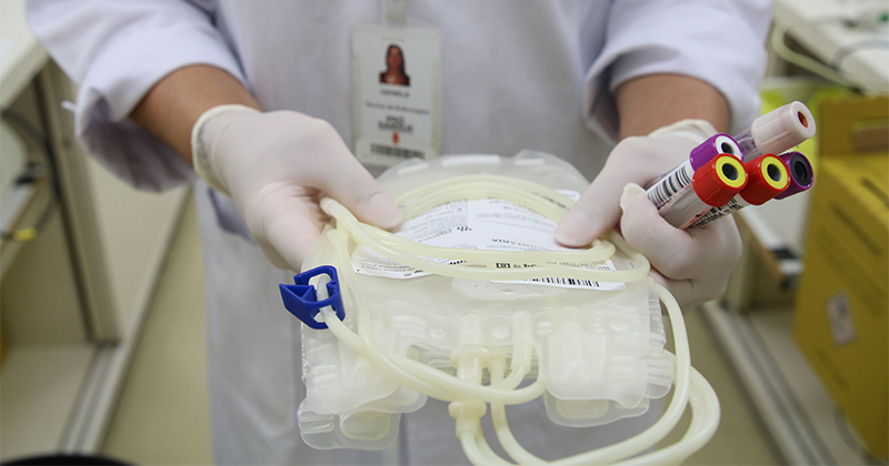 Pessoa vestindo luvas segura bolsas e ampolas de coleta de sangue em ambiente hospitalar