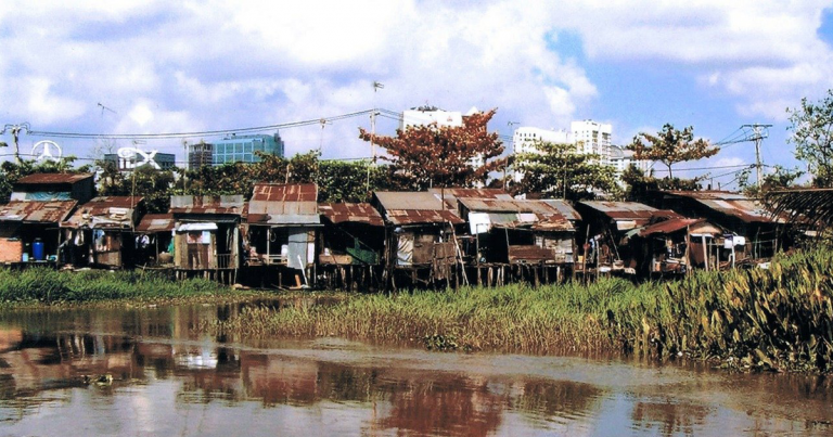 Imagem de uma comunidade periférica situada à beira de um rio, o qual praticamente invade os barracos