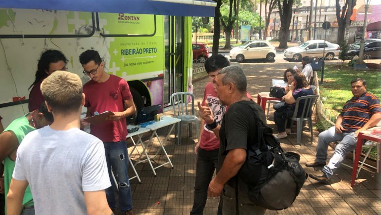 A imagem mostra um atendimento social em um espaço arborizado, possivelmente uma praça. Voluntários, vestindo camisetas vermelhas, interagem com pessoas em situação de vulnerabilidade próximo a um veículo de serviço com identificação de Ribeirão Preto. Um homem de camiseta preta segura um copo enquanto conversa com um dos voluntários. Ao fundo, algumas pessoas estão sentadas em cadeiras de plástico vermelho, aguardando atendimento. A cena transmite um ambiente de apoio comunitário.