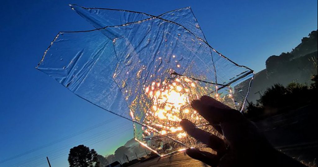 Imagem de uma mão segurando um pedaço de gelo que reflete a luz do sol numa paisagem rural