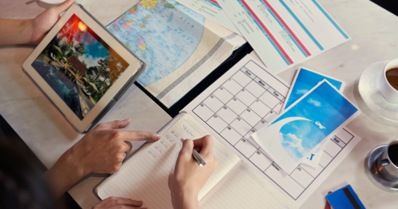 Imagem que mostra, sobre uma mesa, uma mão empunhando uma caneta e escrevendo num caderno de notas tendo ao lado alguém que segura um computador sobre um mapa mundi