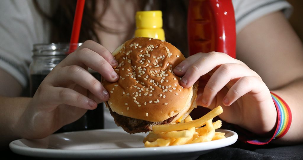 A imagem mostra uma pessoa segurando um hambúrguer com ambas as mãos, prestes a dar uma mordida. O sanduíche tem um pão de gergelim e parece conter carne e queijo. No prato branco à sua frente, há batatas fritas. No fundo, há garrafas de ketchup e mostarda, além de um copo com refrigerante e um canudo vermelho. A pessoa está vestindo uma camiseta clara, e seu braço esquerdo tem uma pulseira colorida com as cores do arco-íris. O foco da imagem está nas mãos e na comida, enquanto o rosto da pessoa não é visível.