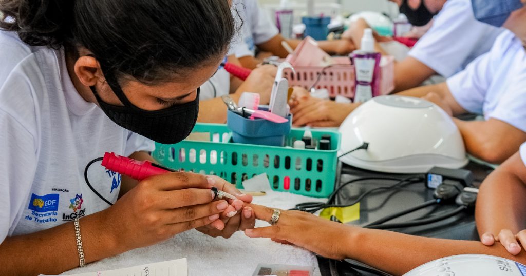 Imagem de uma jovem negra, usando máscara e fazendo manicure numa pessoa cujo rosto não se vê