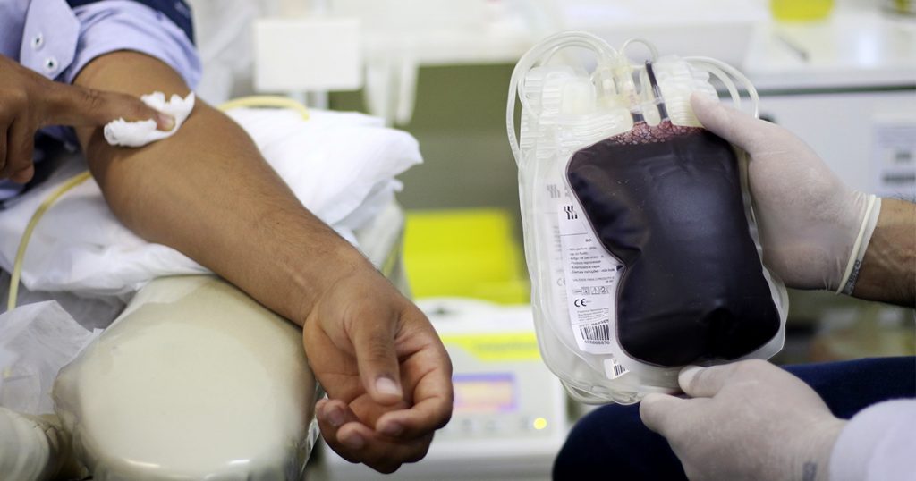 O braço esquerdo de um homem cujo rosto não se vê recebe os cuidados pós doação de sangue, enquanto ao lado alguém com luvas de borracha segura uma bolsa de sangue totalmente cheia