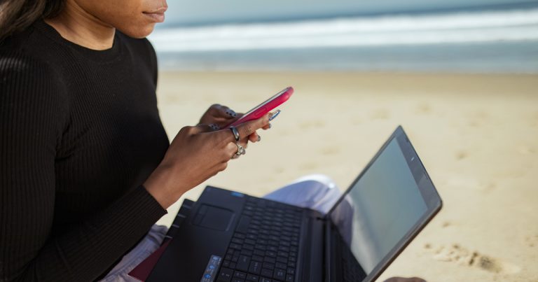 Mulher de camisa preta de manga comprida usando laptop preto e celular de capa vermelha, na praia