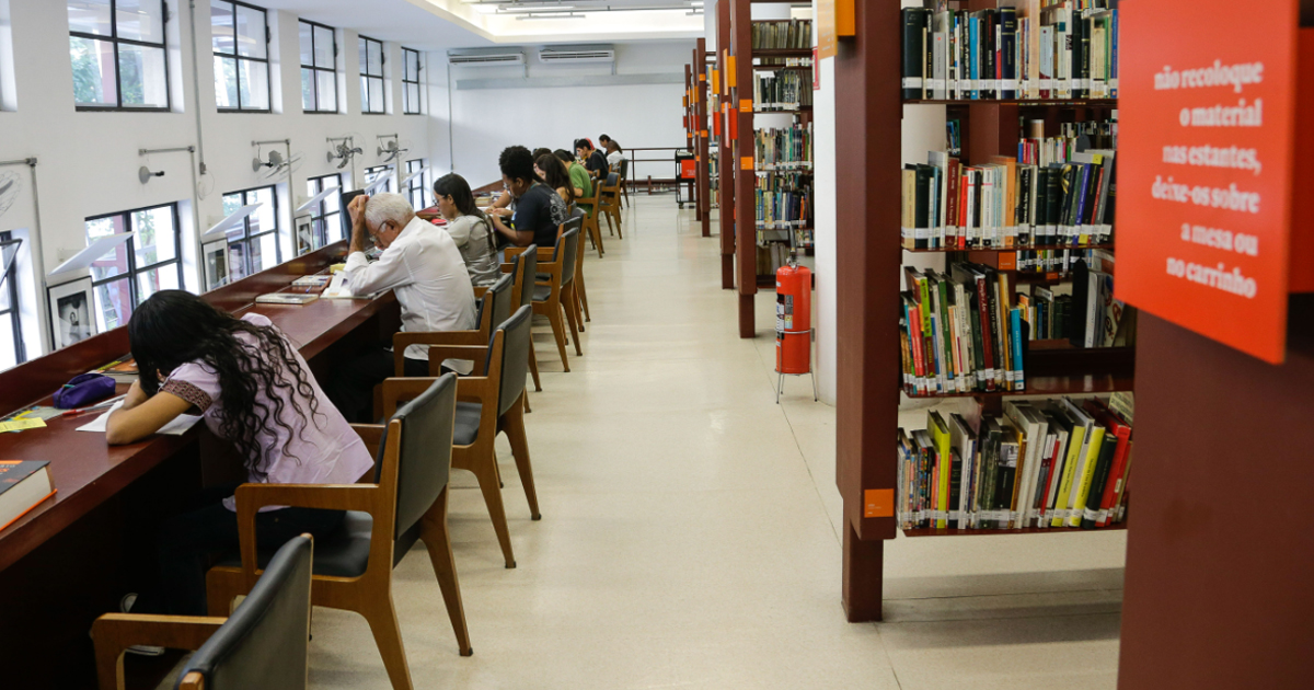 Estudantes sentandos e escrevendo em uma mesa na biblioteca; ao lado estantes de livros