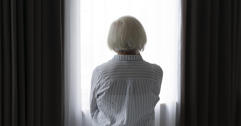 A imagem mostra uma mulher idosa de costas, olhando para uma janela com cortinas escuras.