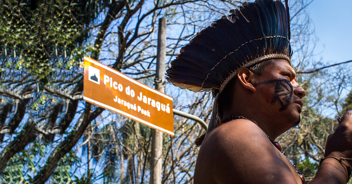 Homem indígena em frente à placa sinalizando a localização do Pico do Jaraguá, na região noroeste de São Paulo. Ele usa um cocar na cabeça e pintura tradicional no rosto.