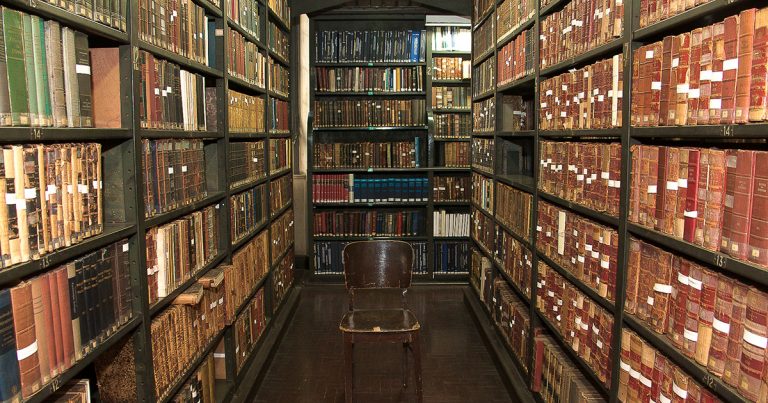 Sala de uma biblioteca, com estantes de livros e uma cadeira no meio do corredor