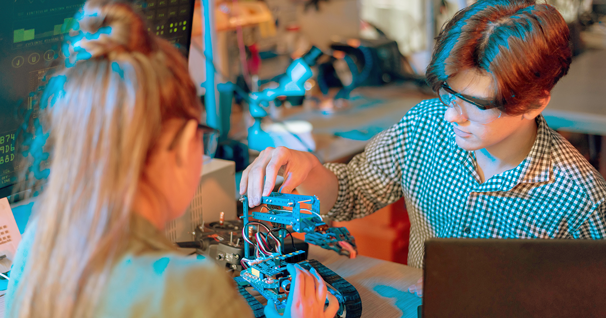 Imagem de um casal de jovens no ato de manusear o protótipo de um robô, no que parece ser uma aula de tecnologia