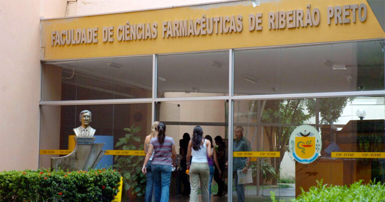 Fachada de um prédio com portas de vidro e uma barra superior em amarelo onde está escrito Faculdade de Ciências Farmacêuticas com duas mulheres de cabelos longos e vestindo calça de camisetas de costas entrando no prédio