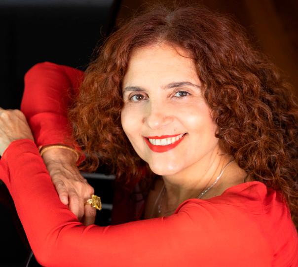 Foto da Beth Amin - mulher branca de cabelos ruivos encaracolados apoiada sobre um piano de cauda.