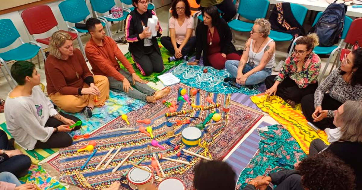 Participantes da edição 2023 do curso de extensão "Arte e Educação com Professores" se sentam sobre tecidos coloridos espalhados pelo chão, em círculo. No centro do círculo há chocalhos, flautas e tamborins.