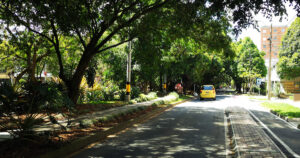 Corredor verde em La Picacha, na cidade de Medelín, na Colômbia. O dia está ensolarado e um carro amarelo se afasta da câmera por uma rua coberta por árvores.