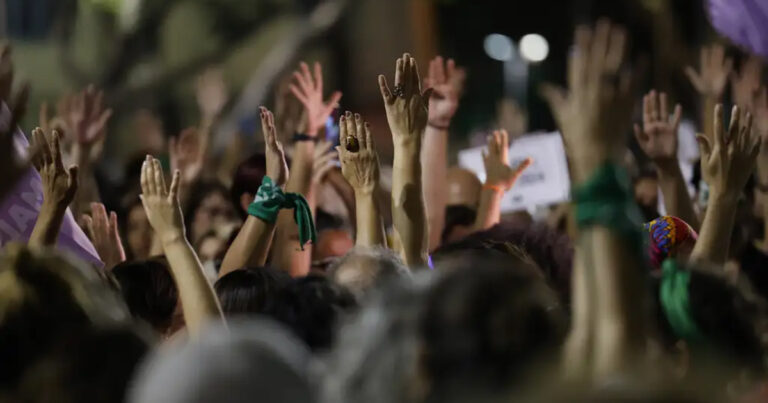 pessoas com as mãos erguidas como se estivessem votando uma proposta em manifestação. Algumas delas carregam um lenço verde amarrado no pulso