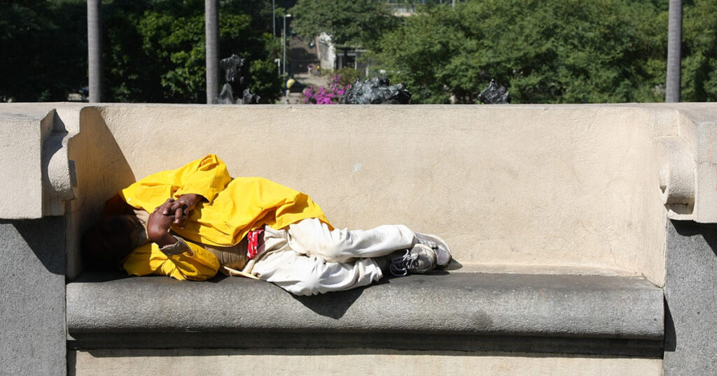 Uma pessoa com casaco amarelo dormindo num banco na rua e com a cabeça coberta