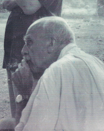 Foto em preto e branco de um homem branco de perfil. É calvo mas tem poucos cabelos brancos. Veste uma camisa branca de mangas curtas. Está fumando um cigarro