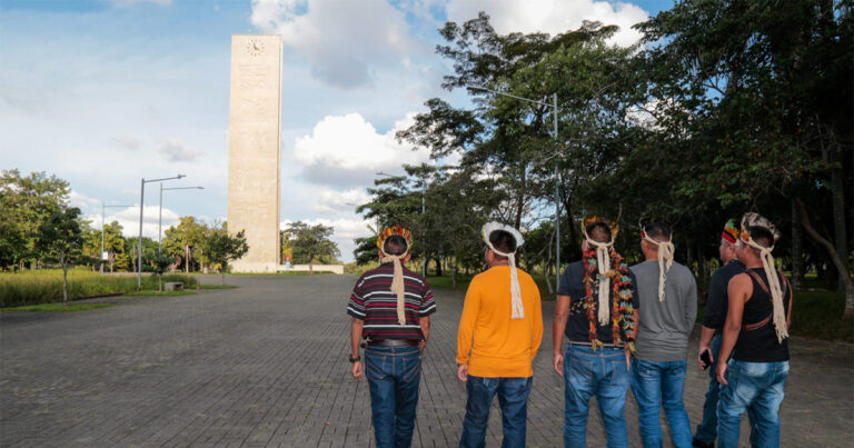 Imagem: Cinco homens indígenas, com trajes de homem branco, caminham pela Praça do Relógio, na Cidade Universitária