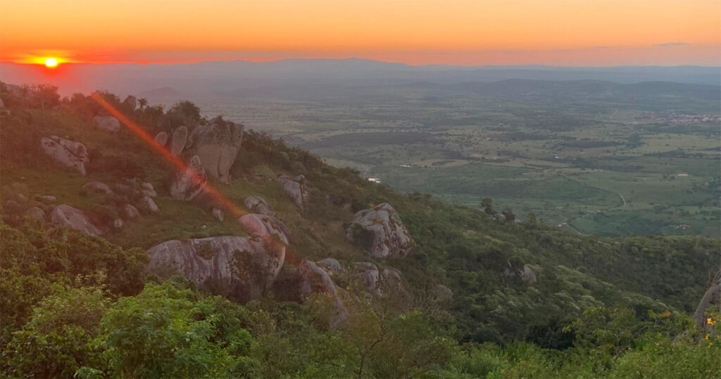 Pôr do sol sobre a paisagem do sertão, com montanhas, árvores e arbustos verdes e encostas rochosas