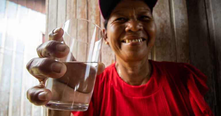 Mulher sorrindo com copo de água na mão. Usa uma camiseta vermelha e um boné