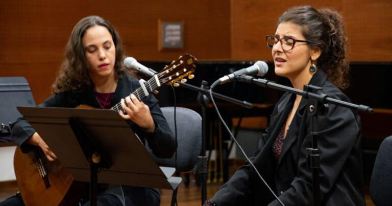 Duas mulheres brancas em um estúdio. A da esquerda toca um violão e ao seu lado, outra mulher usando óculos cantando