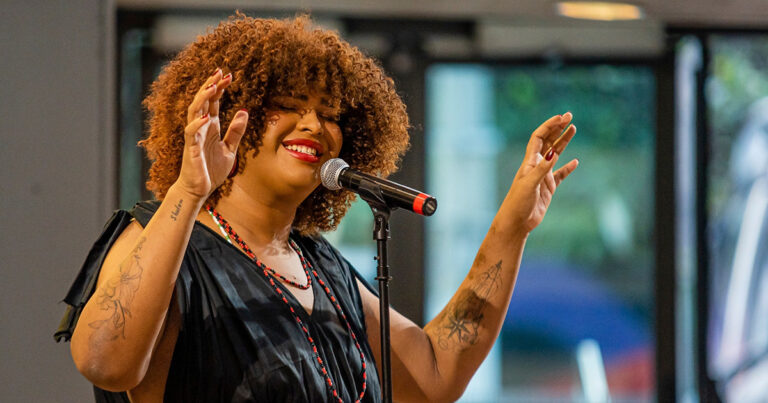 A imagem mostra uma mulher cantando em um microfone. Ela tem cabelo cacheado e volumoso, de cor castanha. Ela usa um vestido preto e colares coloridos. Nos braços, ela tem várias tatuagens visíveis. O fundo da imagem está desfocado, mas parece ser um ambiente interno com janelas grandes.