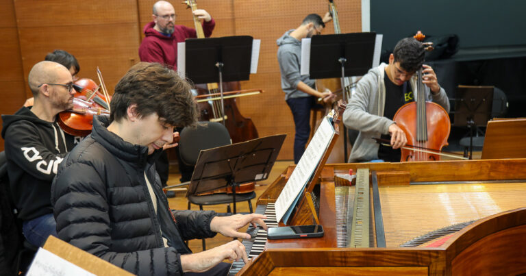 A imagem mostra um grupo de músicos em um ensaio, em ambiente com paredes de madeira. No primeiro plano, há um jovem tocando um piano ou cravo. Ele está usando uma jaqueta preta. À sua direita, um jovem está tocando um violoncelo. Ao fundo, há mais músicos: dois tocando violinos, outros dois, um de moletom vermelho, tocam contrabaixos.