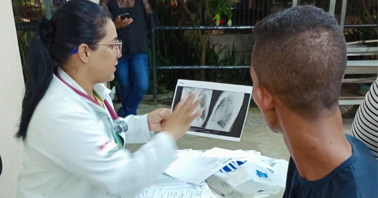 A imagem mostra uma médica, vestida com um jaleco branco e usando um estetoscópio, conversando com um paciente. Ela está segurando e mostrando ao paciente uma radiografia, que parece ser de um tórax. O paciente está de costas, com a cabeça virada para a médica. Ao fundo, há um corrimão, uma pessoa em pé, mexendo no celular, e algumas plantas.