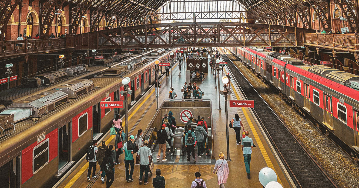 Imagem do alto da estação de trem da Luz, em São Paulo, com as duas linhas de trem na esquerda e na direita e pessoas circulando na área central para embarque