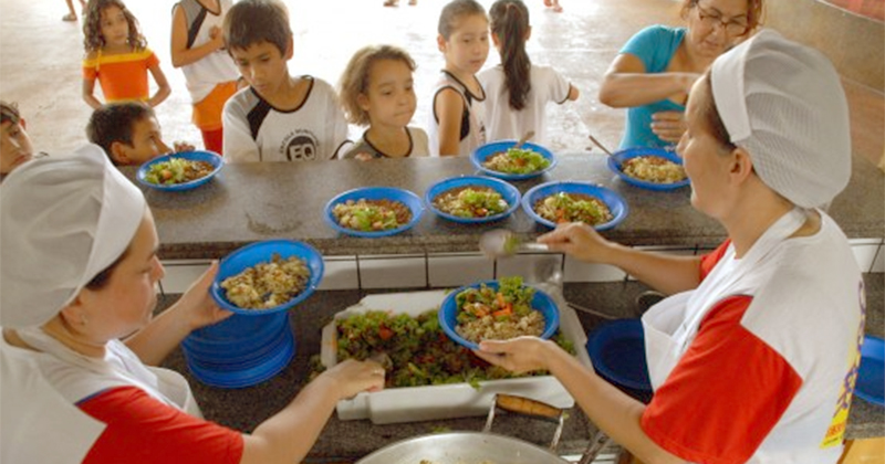 Duas funcionárias distribuindo merenda escolar para um grupo de crianças na escola