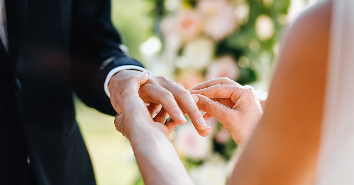 Imagem de duas mãos - uma de um homem, outra de uma mulher -, sendo que a mão da mulher foi captada pela câmera no ato de colocar uma aliança de casamento no dedo anular do homem