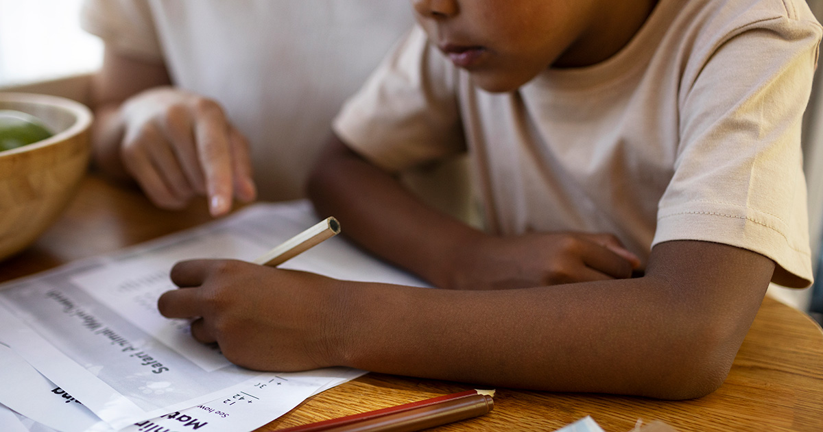 Aluno de escola básica faz dever de casa numa mesa com ajuda da mãe, segura um lápis e escreve numa folha de papel