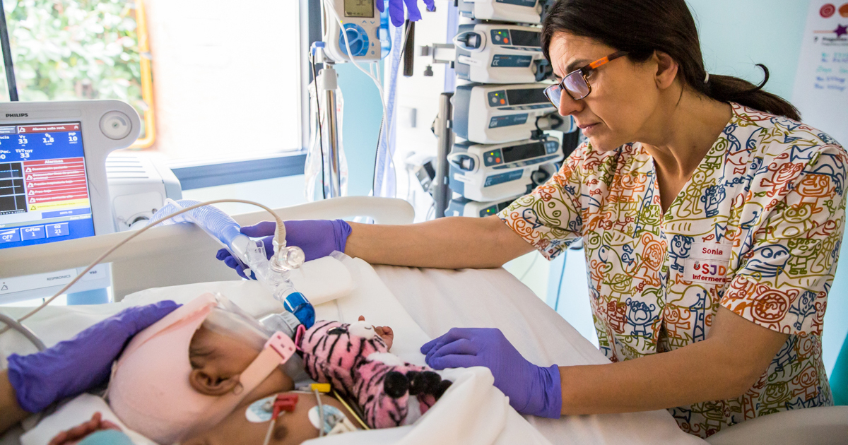 Enfermeira ajusta tubo ligado à máscara de nebulização em bebê deitado em cama de hospital com brinquedo de pelúcia, ao fundo estão os equipamentos hospitalares e uma janela.