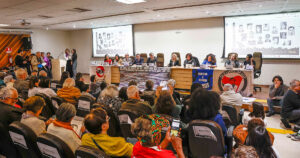 Auditório com pessoas numa mesa em cima de um palco com pessoas assistindo sentadas em cadeiras