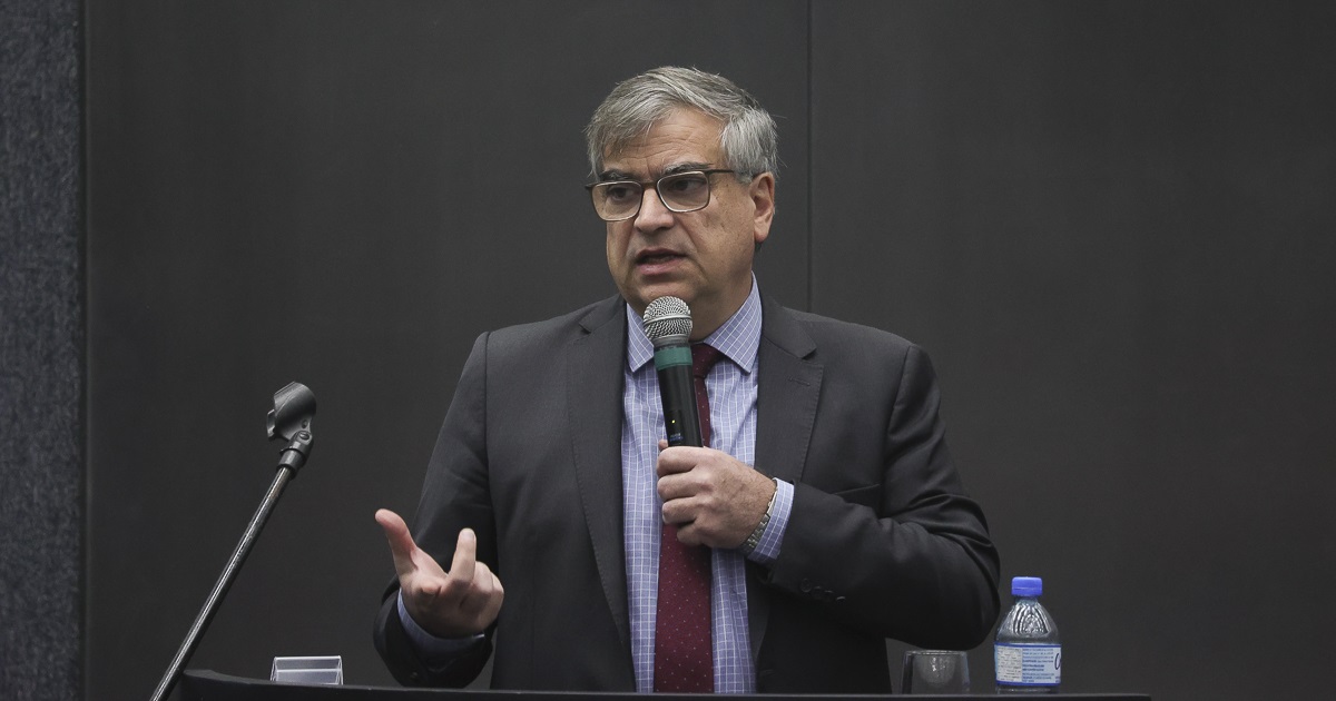 O reitor, homem branco de cabelos grisalhos, vestindo um terno azul escuro, camisa azul clara e gravata bordô, usando óculos, fala com um microfone na mão em um fundo escuro