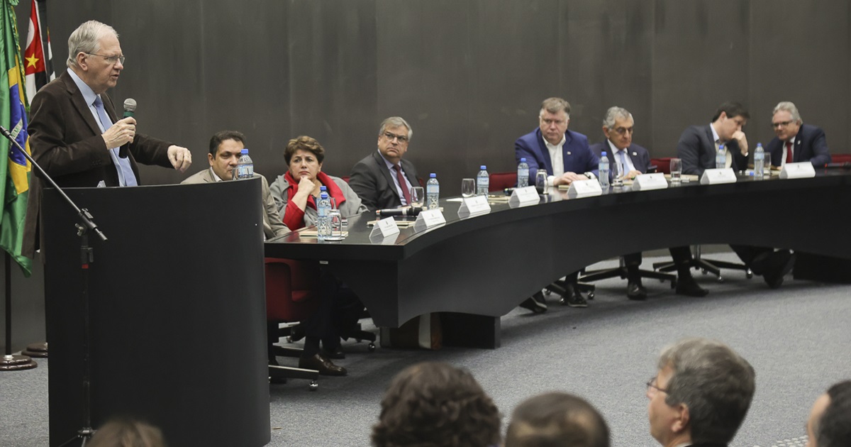 Marco Antonio Zago, homem branco, cabelos brancos, vestindo um terno escuro, está de pé em um púlpito, falando ao microfone. Atrás, bandeiras do Estado de São Paulo e do Brasil. Ao seu lado esquerdo, mesa com 5 pessoas ouvindo. Cabeças de pessoas de costas na plateia aparecem ao pé da foto