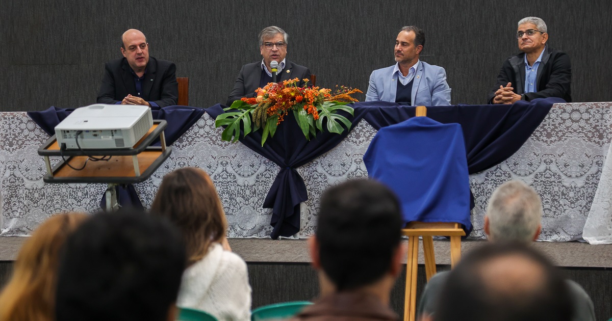 Público de costas assiste a quatro pessoas na mesa que está no palco. Os quatro são homens, e na mesa há uma toalha branca e ornamentos de flores.