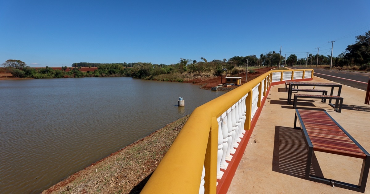 Um lago com um mirante. O mirante contem uma cerca de postes brancos e apoio amarelo, um piso de cimento, um banco de madeira e uma mesa com bancos de madeira. O ceu está muito limpo, azul e ensolarado. Ao fundo do lago, muita vegetação. 