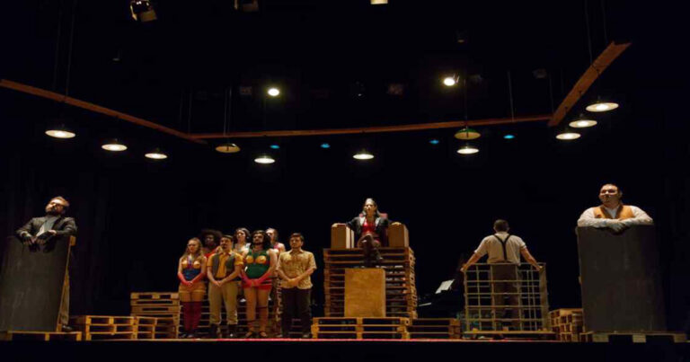 Grupo de atores atuando em peça de teatro sobre palco escuro, mal iluminado.