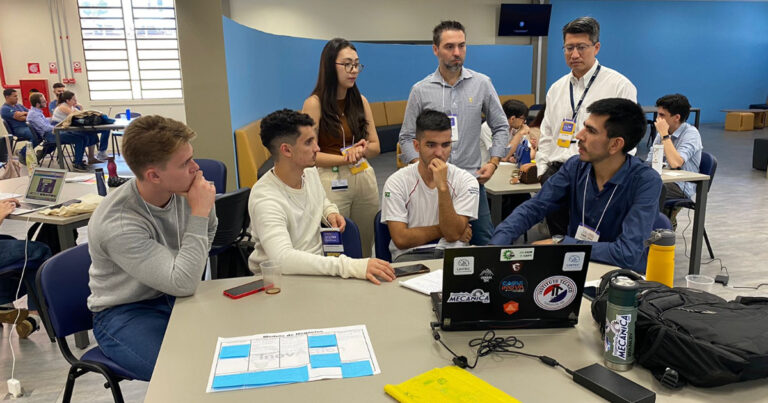 Grupo de quatro estudantes sentados e três em pé em frente a uma mesa com notebook e anotações em papéis