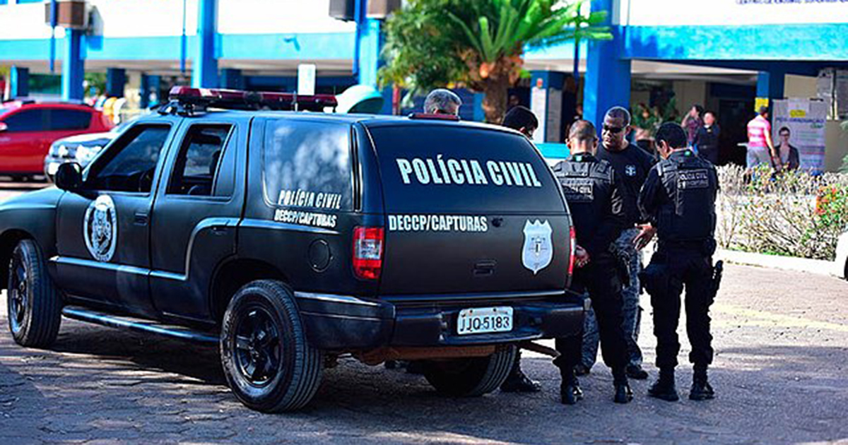 Uma viatura da Polícia Civil estacionada na rua e quatro policiais de colete à prova de balas de pé ao lado dela