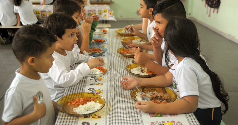 Várias crianças numa mesa de refeitória na escola comendo a merenda escolar num prato com arroz, feijão e carne picada