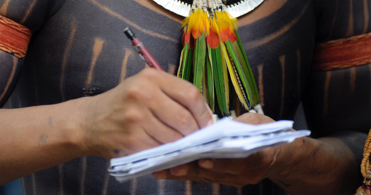 detalhe de homem indígena com pintura, colar e uma caneta e livro nas mãos