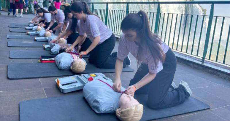 Várias crianças estão sentadas em tapete de EVA simulam ressuscitação cardiopulmonar em bonecos; ao fundo as cachoeiras do Parque Nacional do Iguaçu