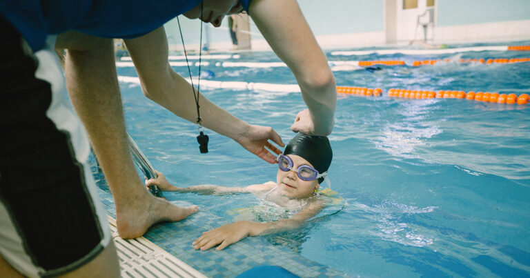 A imagem mostra uma criança em uma piscina, usando uma touca de natação preta e óculos de natação roxos. Está na borda da piscina, com as mãos apoiadas na borda. Um adulto, que parece ser um instrutor de natação, está ao lado dela, ajustando a touca da criança. O instrutor usa um apito pendurado no pescoço. No fundo, há boias laranjas que delimitam as raias da piscina