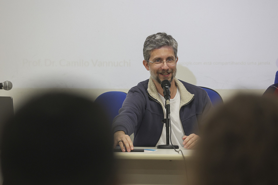 homem branco, barba, bigode e cabelos curtos e grisalhos, óculos de aro fino. Usa uma jaqueta de moletom sobre uma camiseta branca