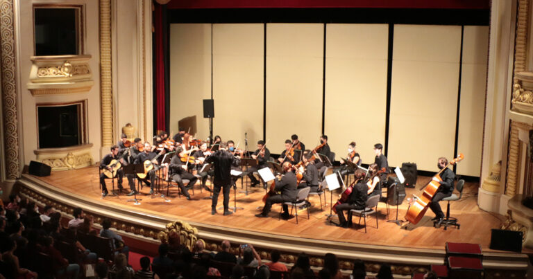 A imagem mostra uma orquestra se apresentando em um teatro. O palco é grande e está ocupado por vários músicos tocando instrumentos de cordas, como violinos, violoncelos e contrabaixos. O maestro está de pé no centro, conduzindo a orquestra. O teatro é elegante, com detalhes ornamentados nas paredes e varandas laterais