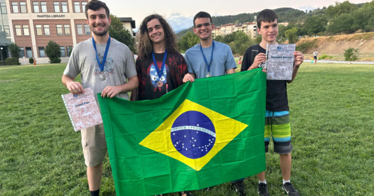 Quatro alunos mostram diplomas e medalhas enquanto seguram a bandeira do Brasil em um gramado ao ar livre; ao fundo um edifício