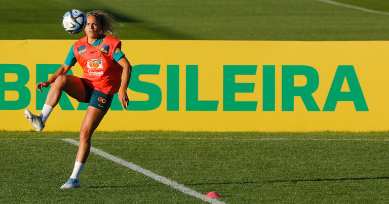 Na imagem, a zagueira Mônica Alves da seleção brasileira aparece fazendo uma embaixadinha em um campo de futebol. Mônica tem cabelos loiros presos em um rabo de cavalo e usa um colete vermelho, com o símbolo da marca Guaraná. No fundo do campo, há uma faixa amarela com a palavra Brasileira, escrita na cor verde e com letras maiúsculas